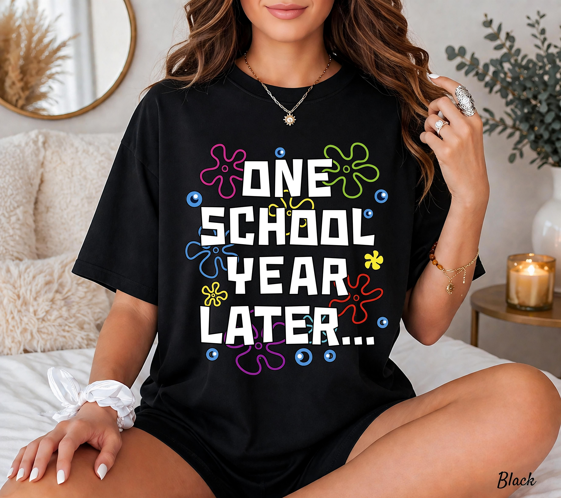 Unisex black t-shirt featuring a colorful retro graphic with white block text reading 'ONE SCHOOL YEAR LATER...'. The background is decorated with colorful cartoon flower shapes and bubbles.